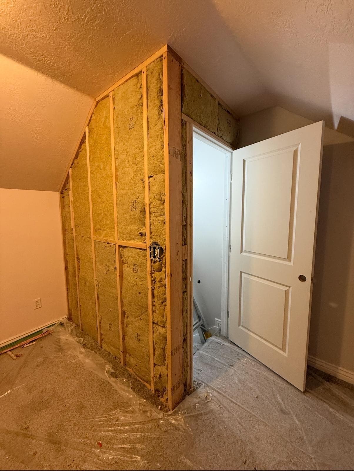 Attic knee wall framed and insulated, the door set in its opening — the stage before drywall closes the corner.